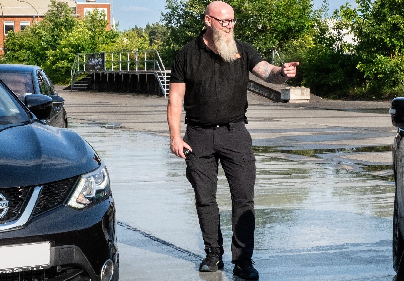 Mann mit Bart und Brille, in schwarzer Kleidung, steht zwischen Autos und gestikuliert.